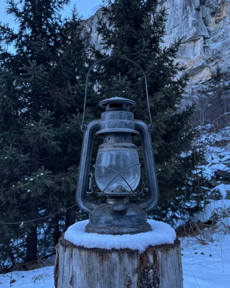 Alte eiserne Öllaterne mit gesprungenem Glas auf einem verschneiten Baumstumpf vor Fichten und hellen Kalkfelsen in der Winterdämmerung