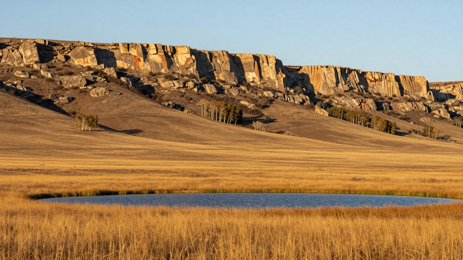 Goldenes Grasland der nördlichen Plains vor der steilen Felswand der Rocky Mountains im späten Nachmittagslicht, ein kleiner See im Vordergrund