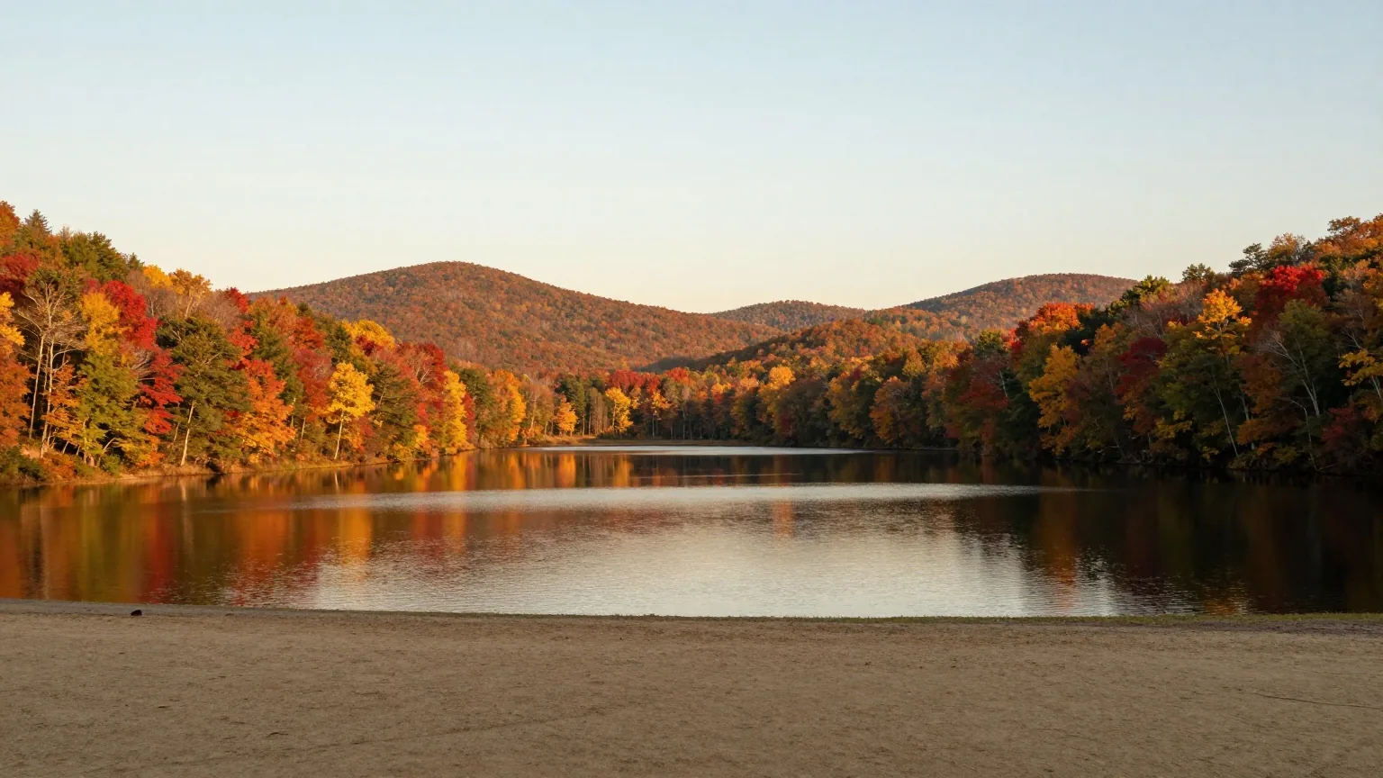 Langer, schmaler See zwischen sanften, bewaldeten Hügeln im Herbst, Laubbäume in Rot und Gold, ruhiges Wasser im Licht der tiefen Nachmittagssonne