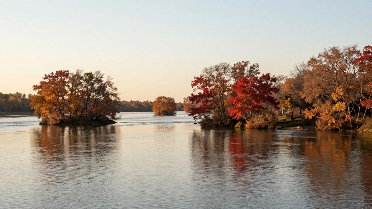 Breiter Fluss mit bewaldeten Ufern und kleinen Inseln im Herbst, Laubbäume in Rot und Gold, tiefstehende Nachmittagssonne über dem Wasser