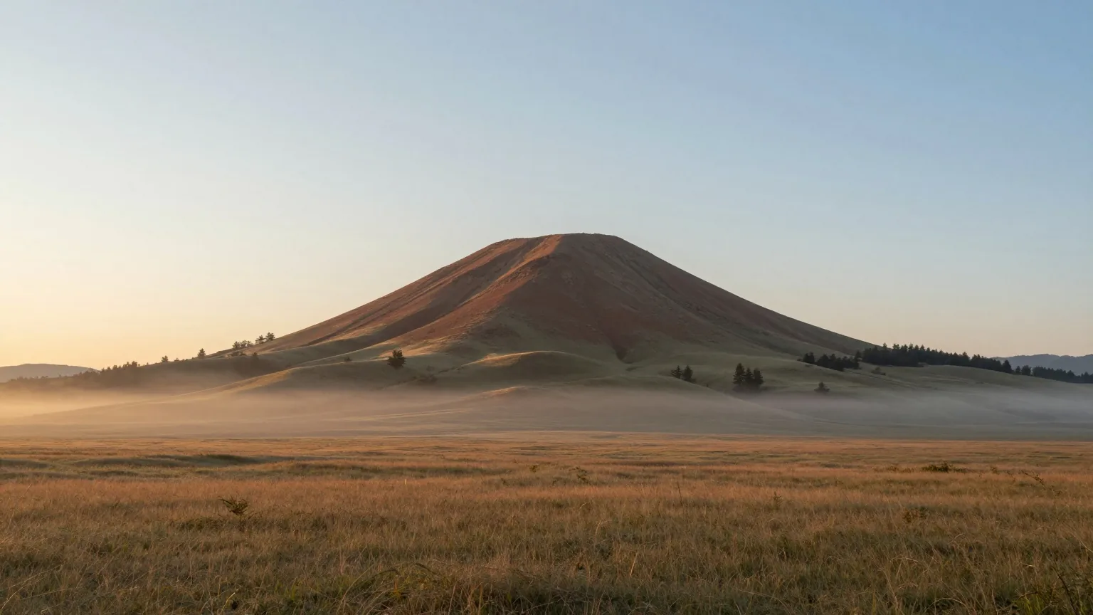 Einzelner kegelförmiger Berg erhebt sich in der Morgendämmerung aus einer weiten Graslandschaft, über dem Horizont leuchtet ein schmaler Streifen Licht