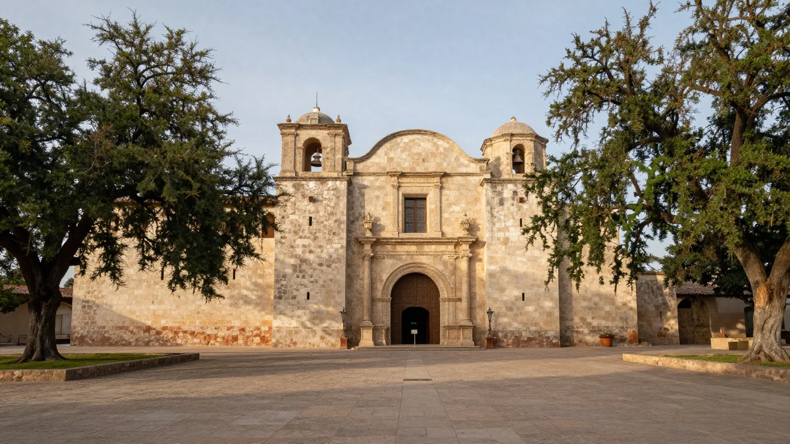 Hellgraue Fassade einer Missionskirche aus Kalkstein mit geschwungenem Giebel und Rundbogenportal, davor ein leerer Platz mit Eichen im warmen Nachmittagslicht