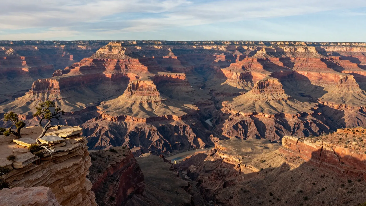 Blick vom Südrand über gestaffelte rote und hellbraune Felswände des Grand Canyon, tief unten ein schmales Flussband, Wacholder am Rand im Nachmittagslicht