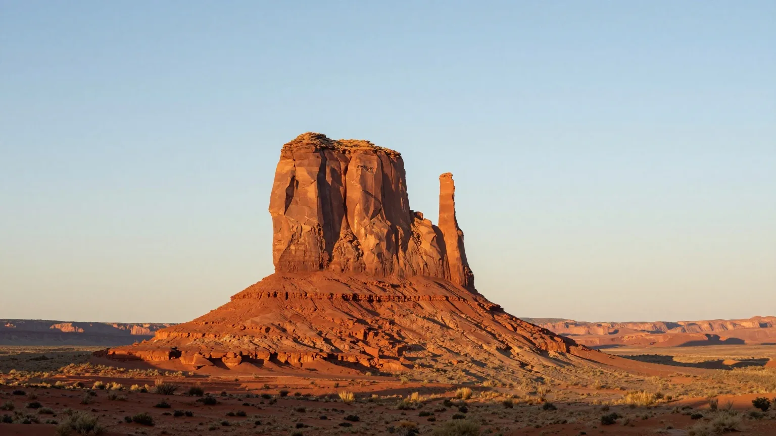 Zwei rote Sandsteinbuttes mit schmalen Felsfingern über einer weiten Wüstenebene mit Sand und Büschen im warmen Licht der tief stehenden Sonne