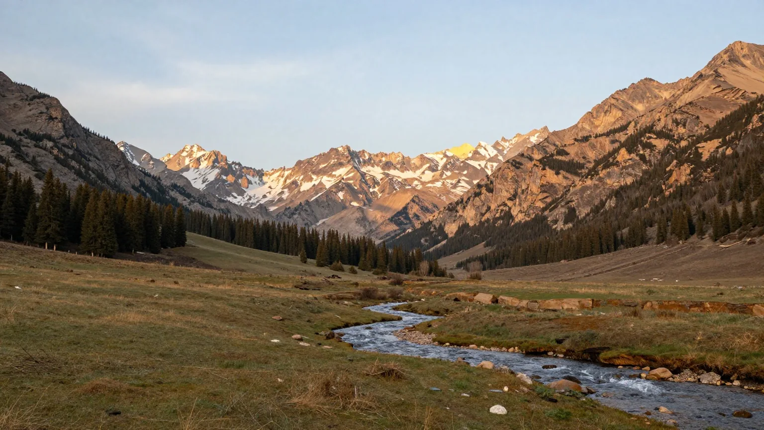 Hohe, teils verschneite Bergkette über einem weiten Hochtal mit Gras, Nadelwald und einem gewundenen Bach im Licht der späten Nachmittagssonne