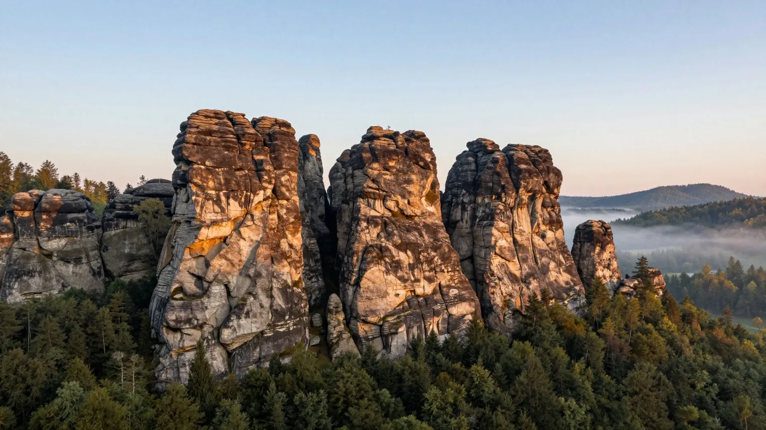 Sandsteinfelsen über einem bewaldeten Tal im Elbsandsteingebirge, die Felstürme leuchten im warmen Licht der späten Nachmittagssonne