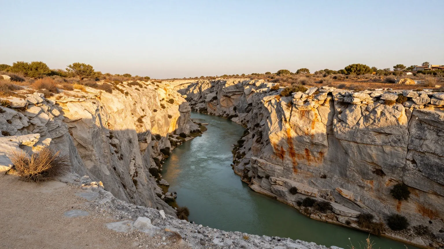Tiefer Kalksteincanyon, durch den sich ein grüner Fluss zwischen hellgrauen Felswänden windet, oben am Rand trockenes Buschwerk im Licht der späten Nachmittagssonne