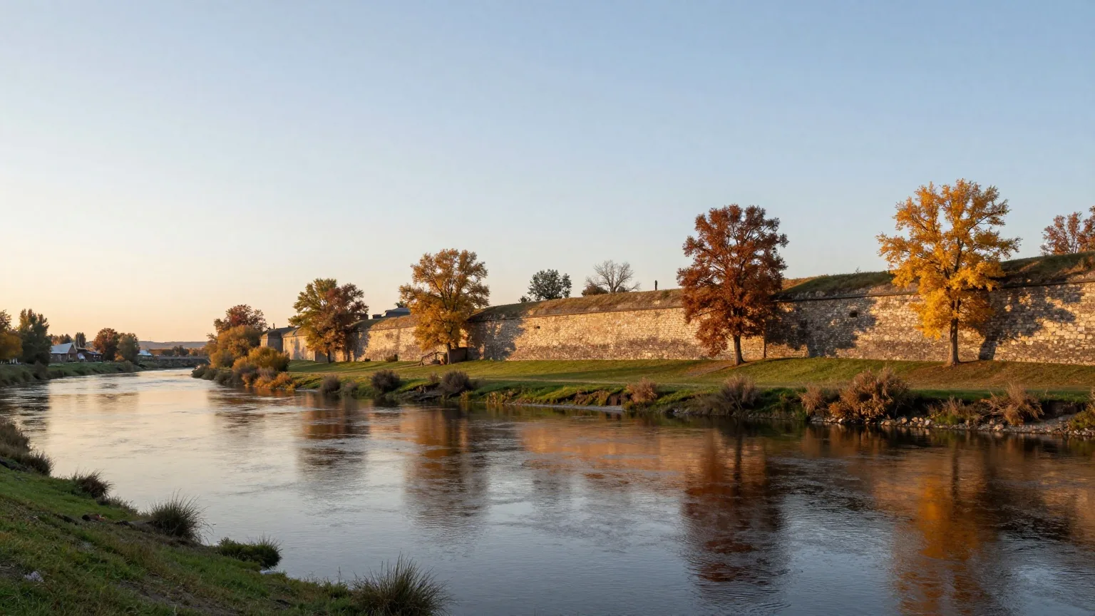 Breiter Fluss im Abendlicht, am Ufer die niedrigen Reste einer alten Steinmauer und Bäume in Herbstfarben