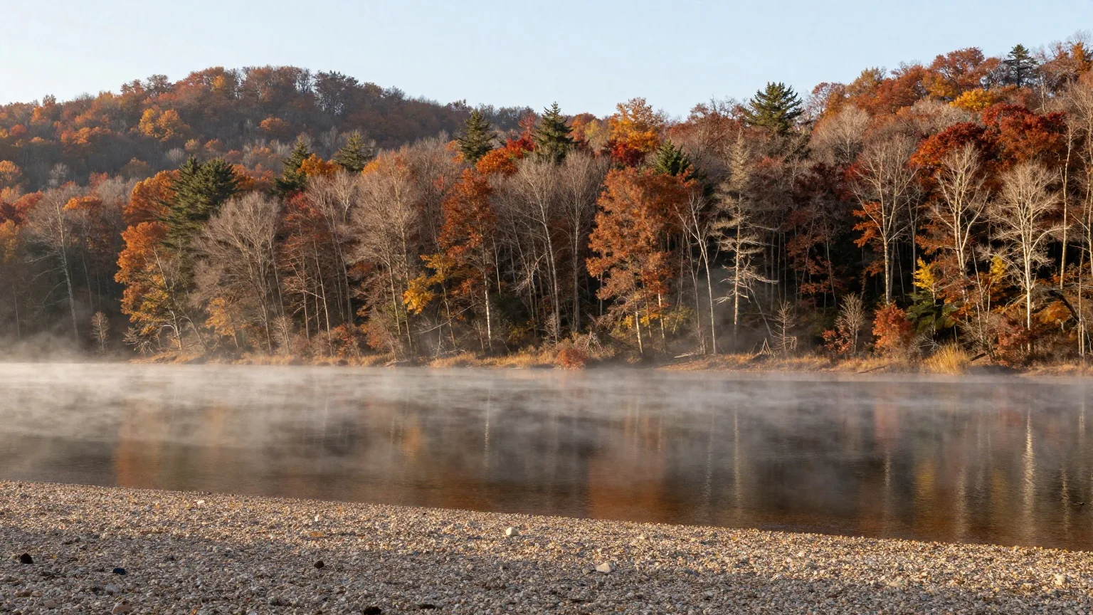 Stiller See zwischen dicht bewaldeten Hügeln mit ersten Herbstfarben, Dunst über dem Wasser im Licht der späten Nachmittagssonne