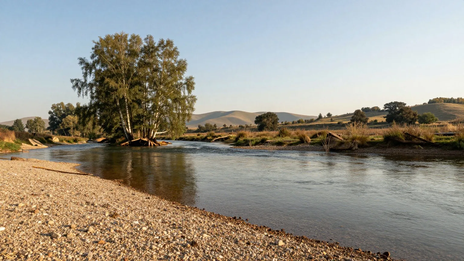 Ruhiger klarer Fluss in einem grünen Tal mit Kiesufer, am anderen Ufer hohe Birken und Eichen, dahinter dunstige bewaldete Hügel im warmen Nachmittagslicht