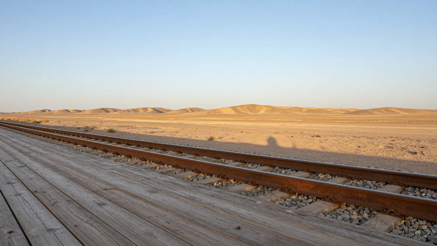 Leerer Bahnsteig aus verwitterten Holzbohlen neben einem einzelnen rostigen Gleis in der Wüste, am Horizont flache Sandhügel im Nachmittagslicht